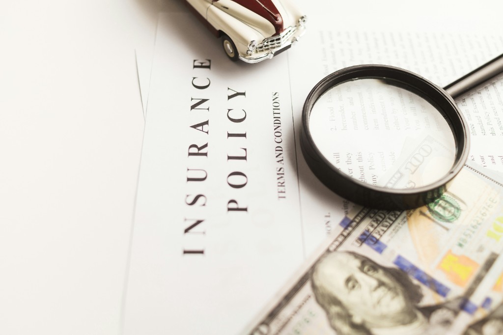 Insurance policy paperwork with magnifying glass, model car, and cash on a white surface
